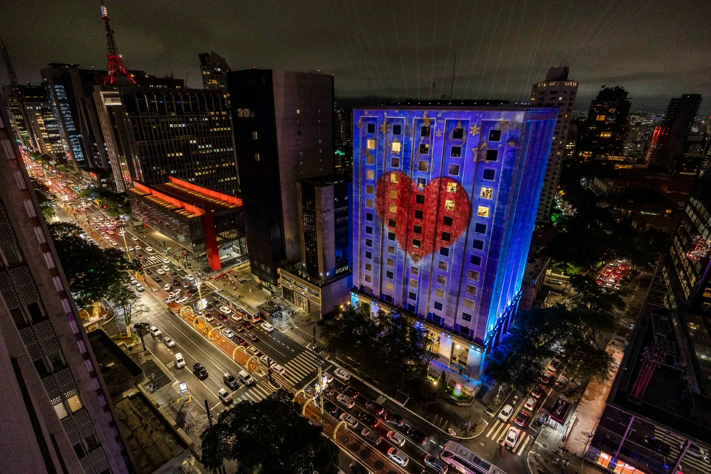Vista aérea noturna da Avenida Paulista: a sede da Bradesco Seguros iluminada em azul, com um laço vermelho projetado ao centro da fachada