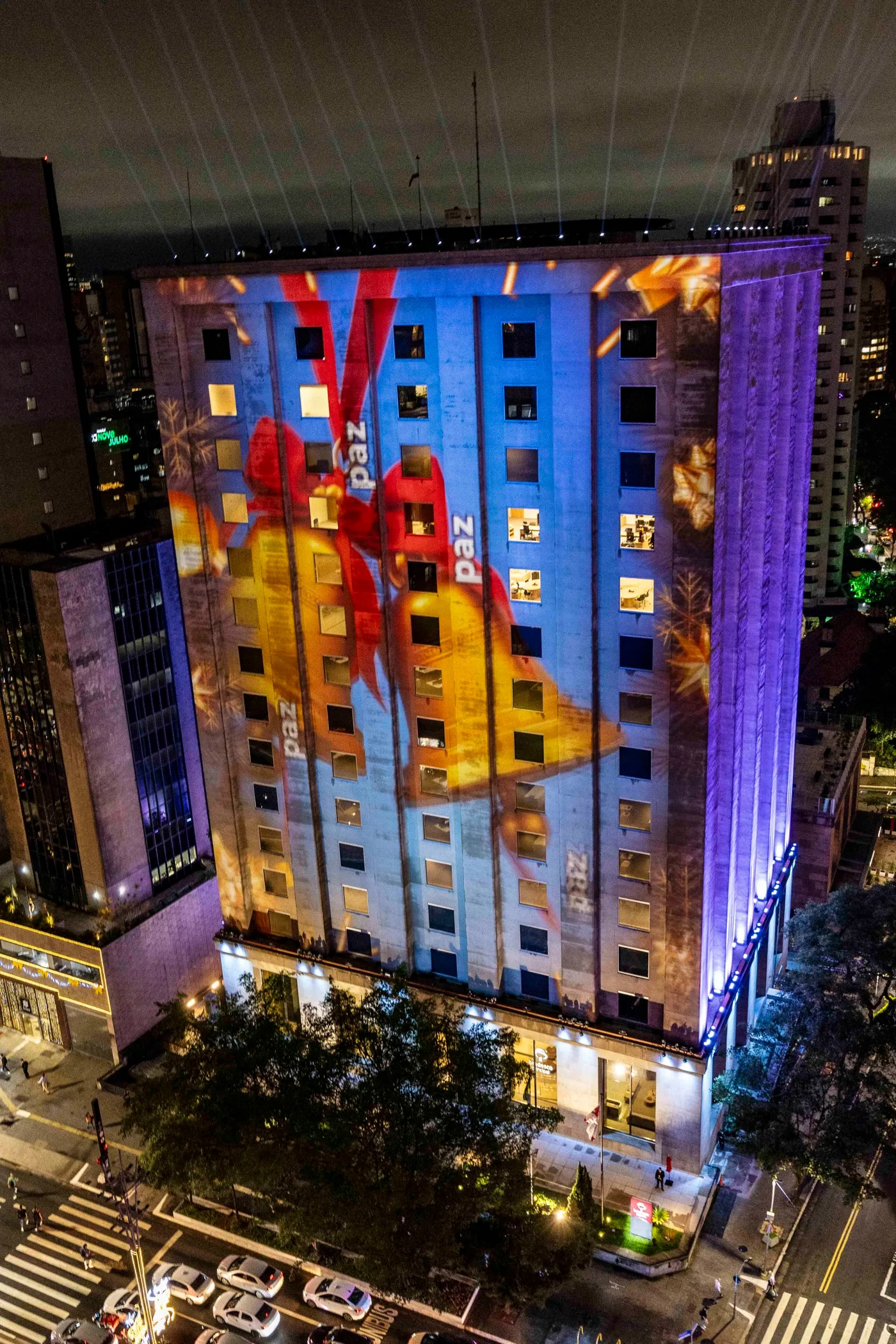 Enquadramento vertical da fachada: um grande laço vermelho projetado sobre o edifício, com a palavra paz repetida ao longo dos andares